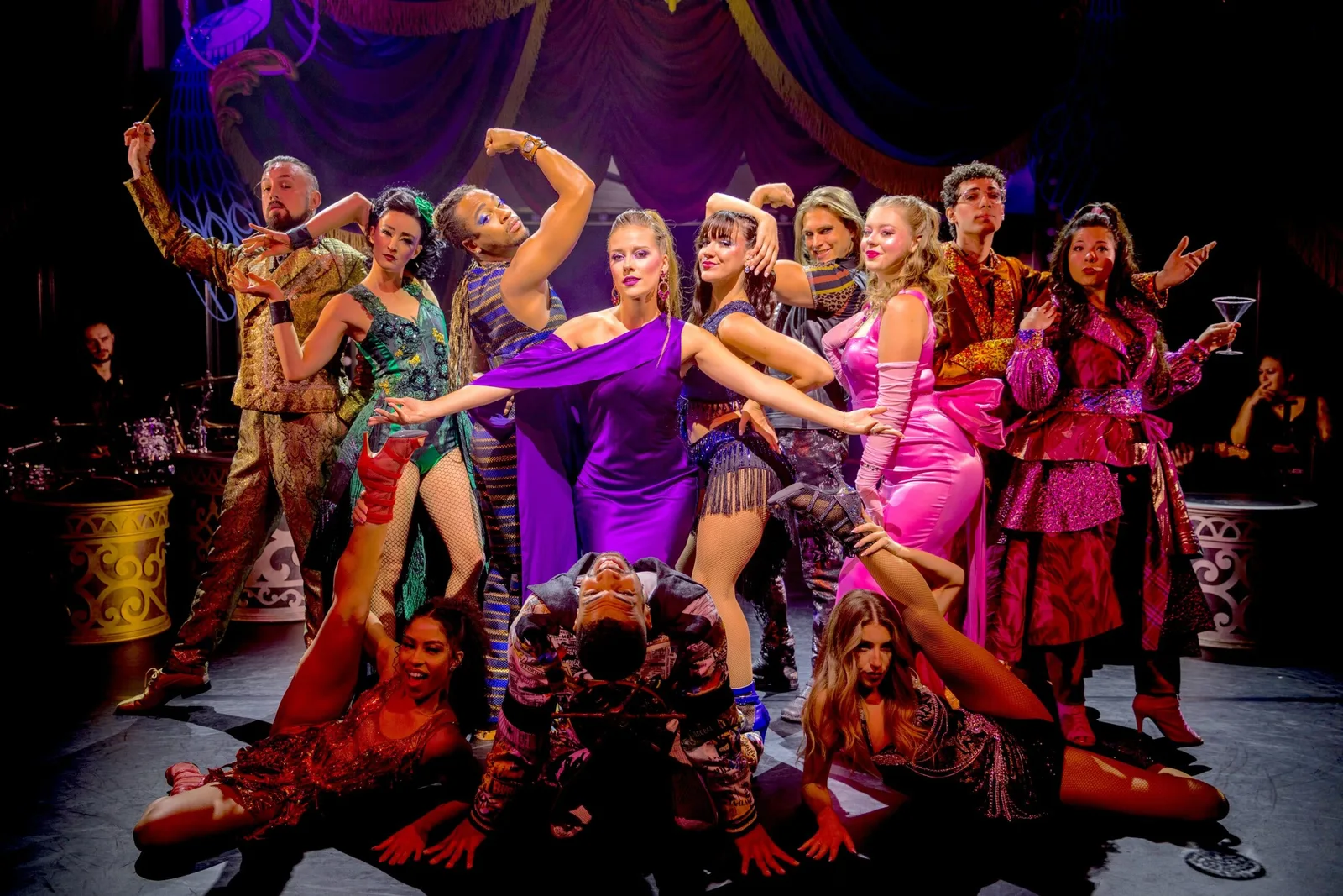 A group of performers in colorful costumes pose dramatically on stage under purple lighting.