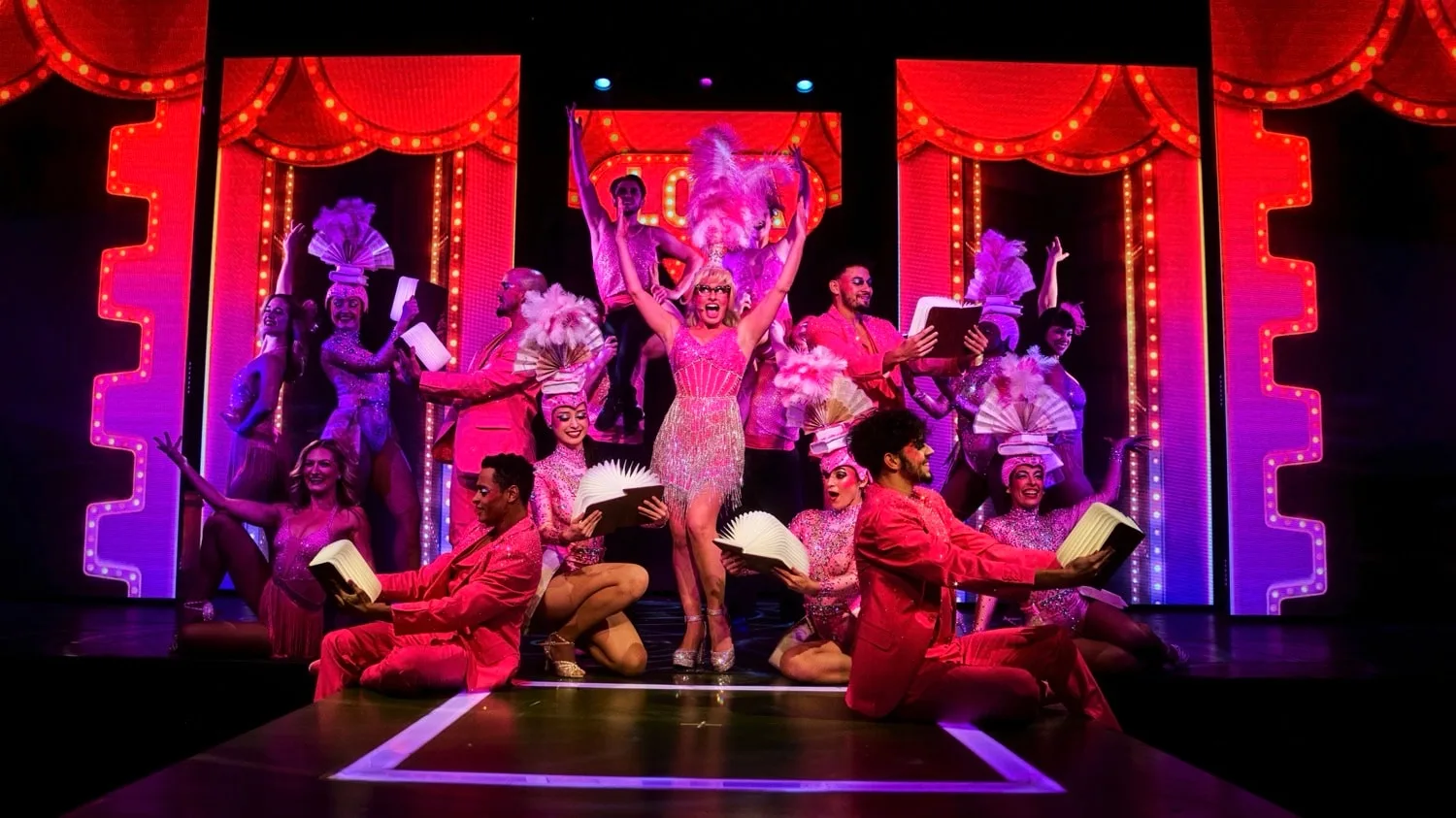 A colorful stage performance featuring dancers in vibrant costumes with feathered headpieces and fans, set against illuminated red and purple backgrounds.