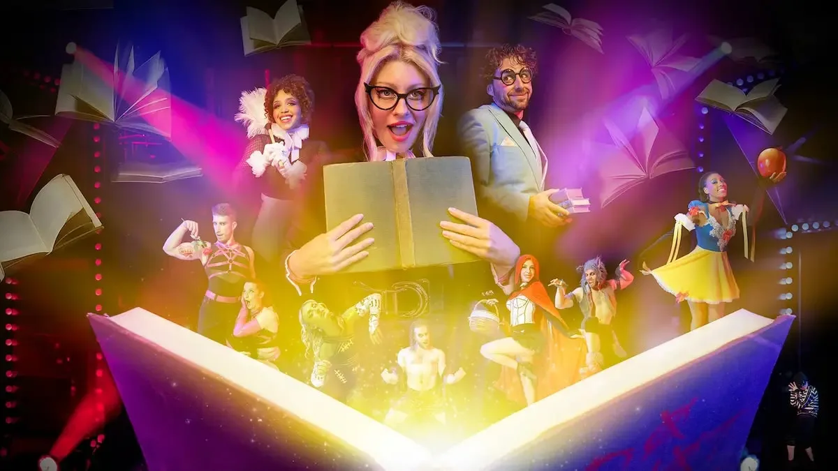 A group of performers in colorful costumes pose on a stage with a large open book prop emitting light and floating pages in the background.