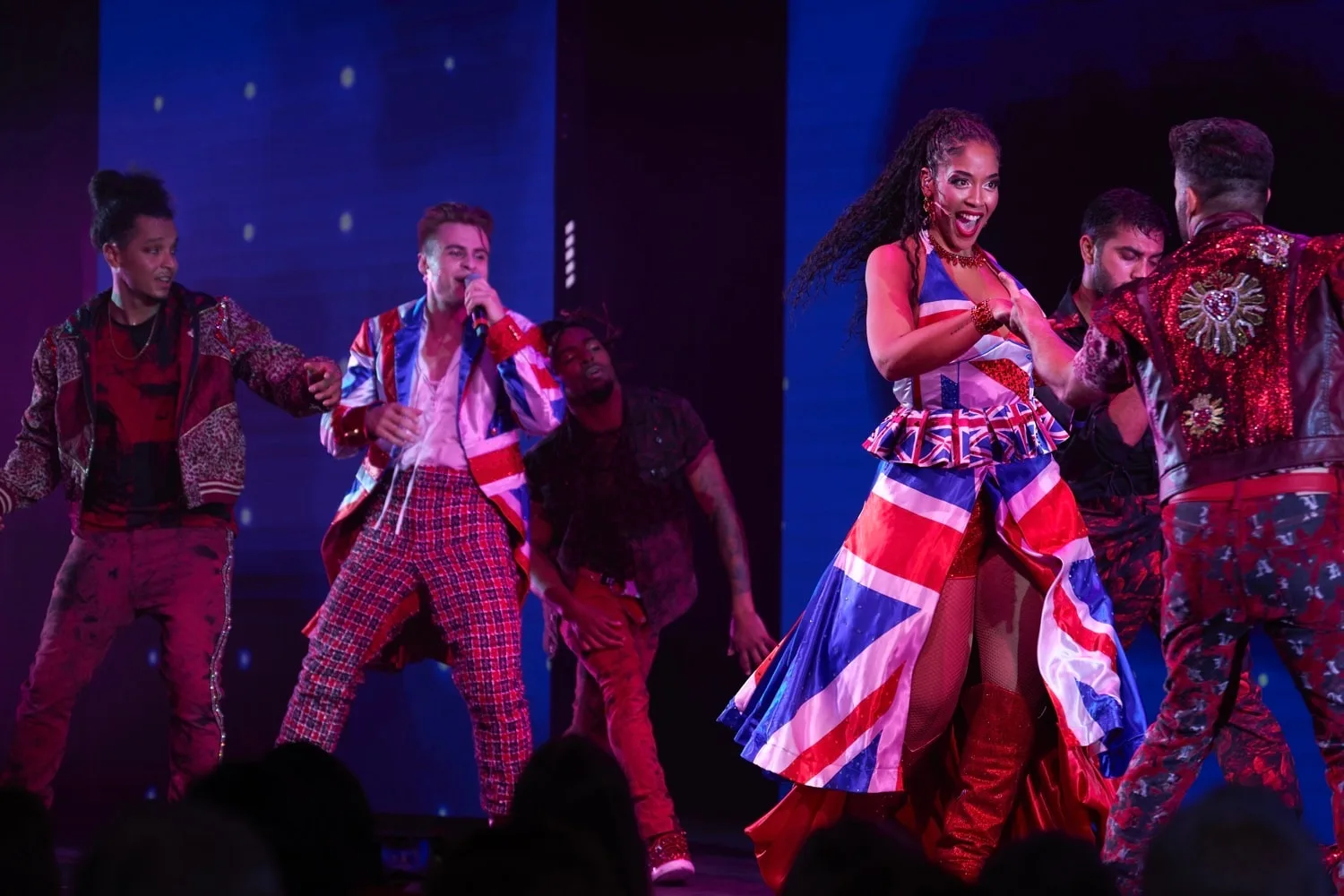 A group of performers are dancing on stage, with one woman prominently wearing a dress featuring the Union Jack flag design.