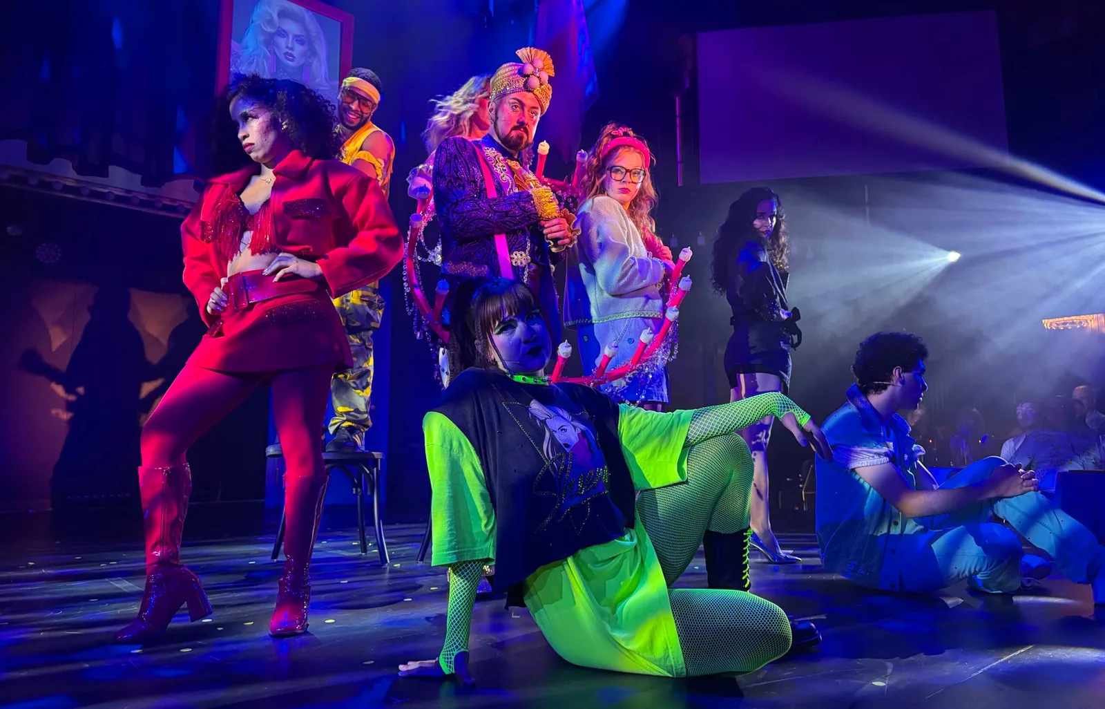 Stage performers in colorful and eclectic costumes pose dramatically under vibrant lighting during a theatrical production.