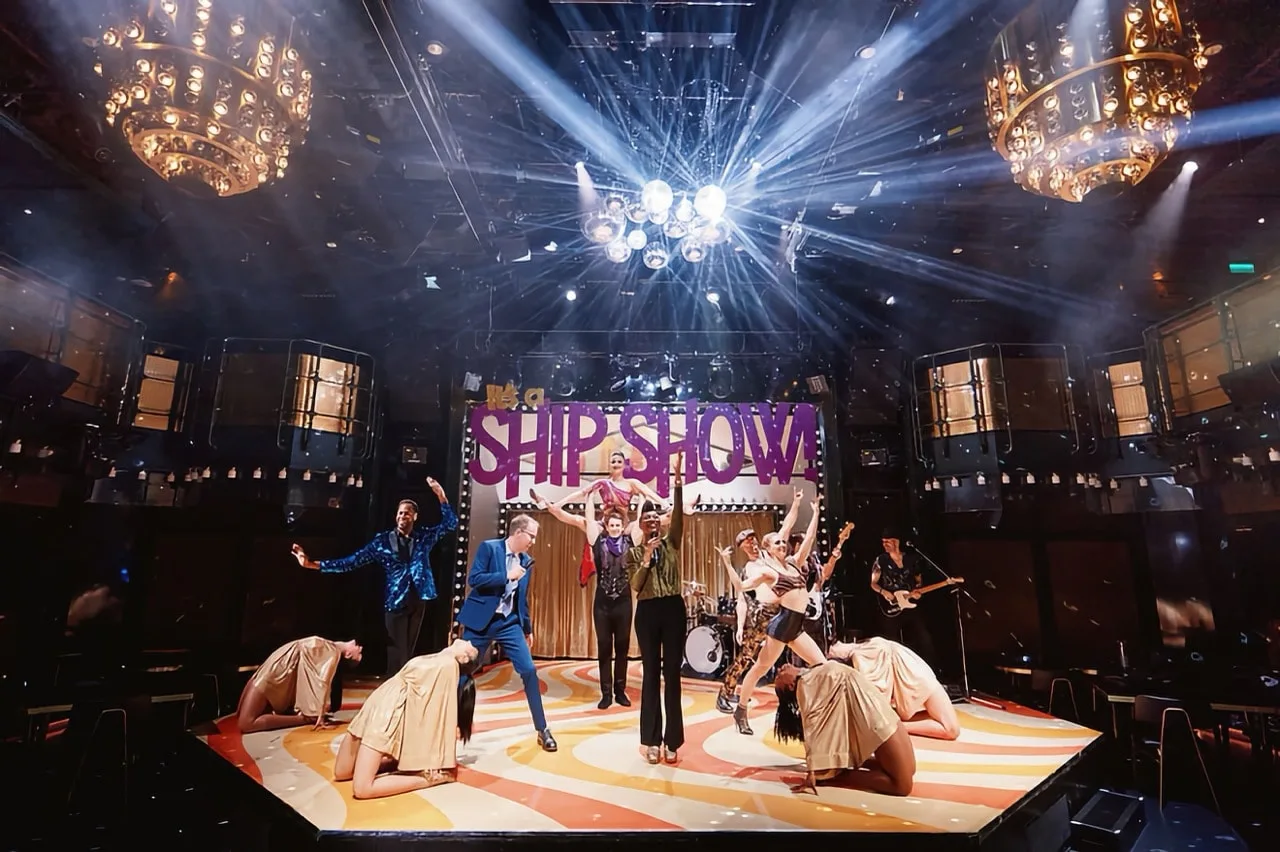 Stage performance of people dancing and singing under chandeliers with a large "SHIP SHOW" sign in the background.