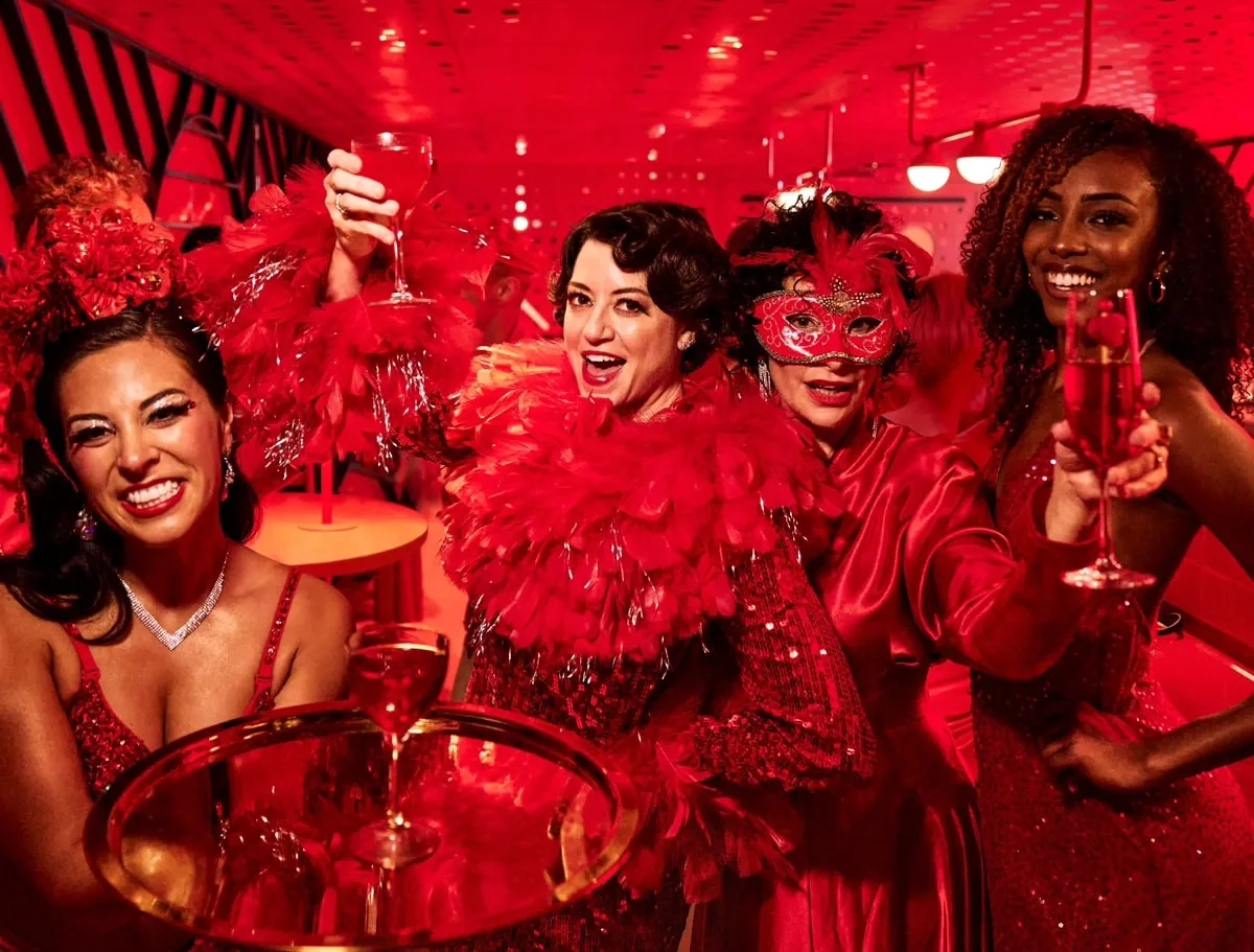 Three women dressed in elaborate red costumes and makeup are seated at a round table inside a red-lit room.