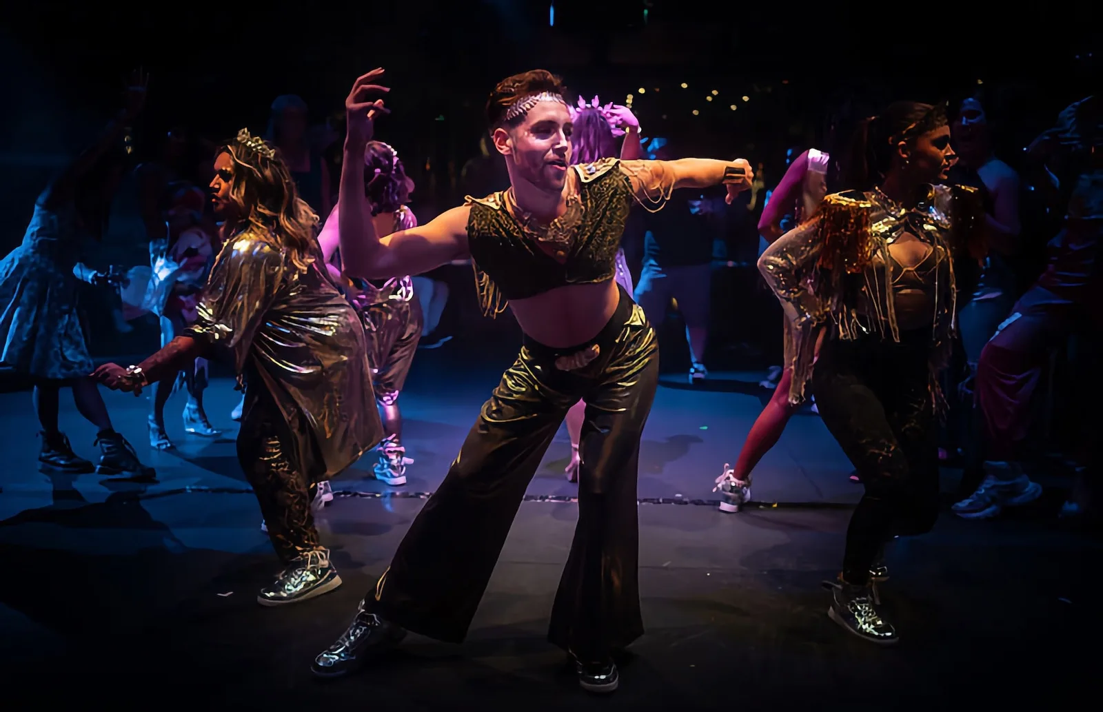 A group of performers dressed in shiny, metallic outfits dance on a dimly lit stage.