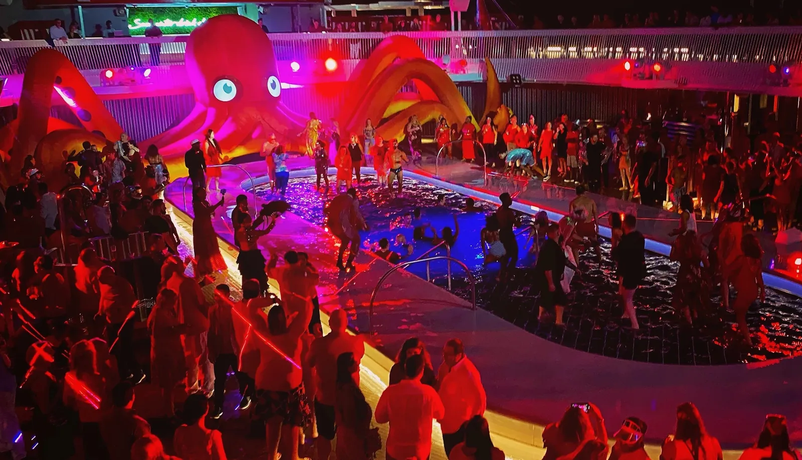 Crowded nighttime pool deck on a cruise ship with a large inflatable red octopus and colorful lights.