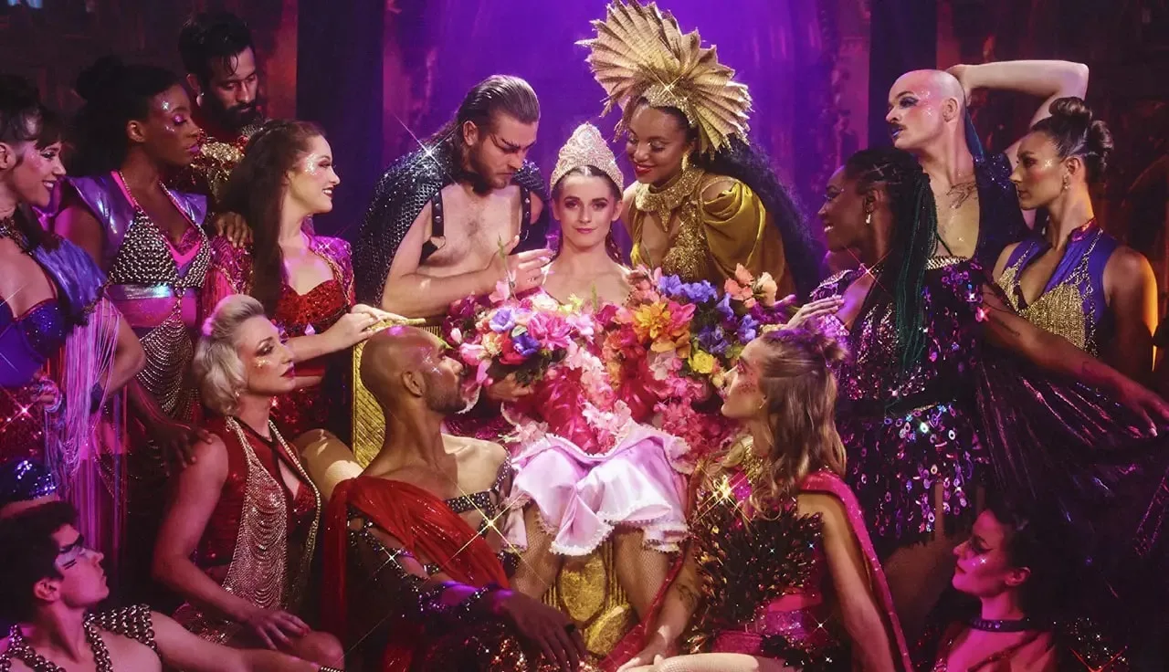 A group of elaborate costumed performers surrounds a woman seated on a throne, adorned with colorful flowers and theatrical outfits.