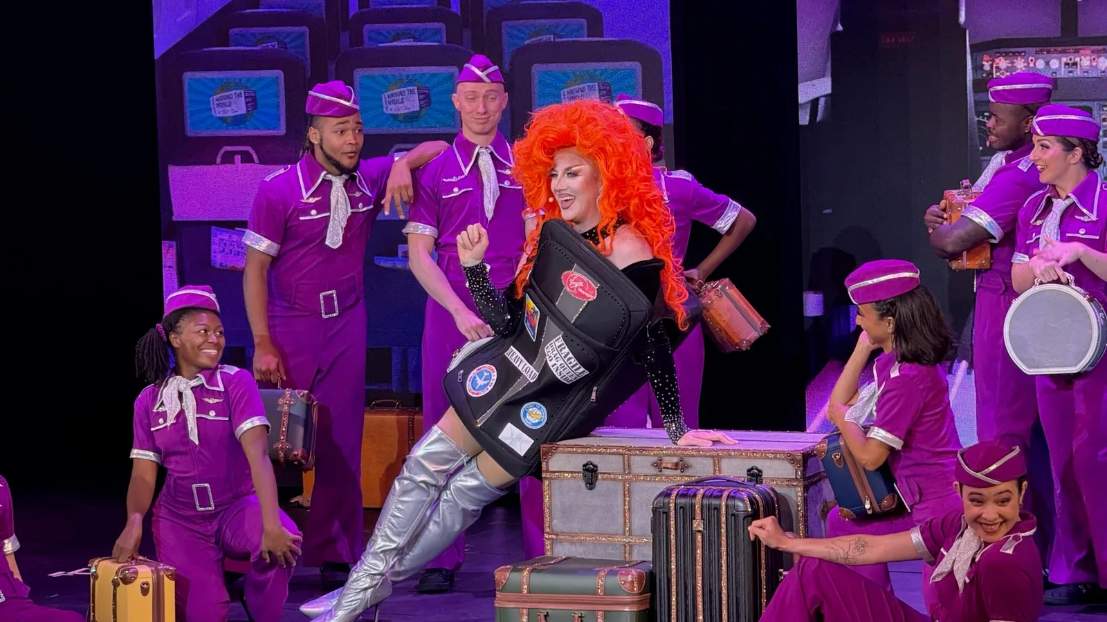 A group of performers dressed in purple airline-style uniforms and a performer with bright orange hair in black attire are on stage with vintage suitcases and luggage.