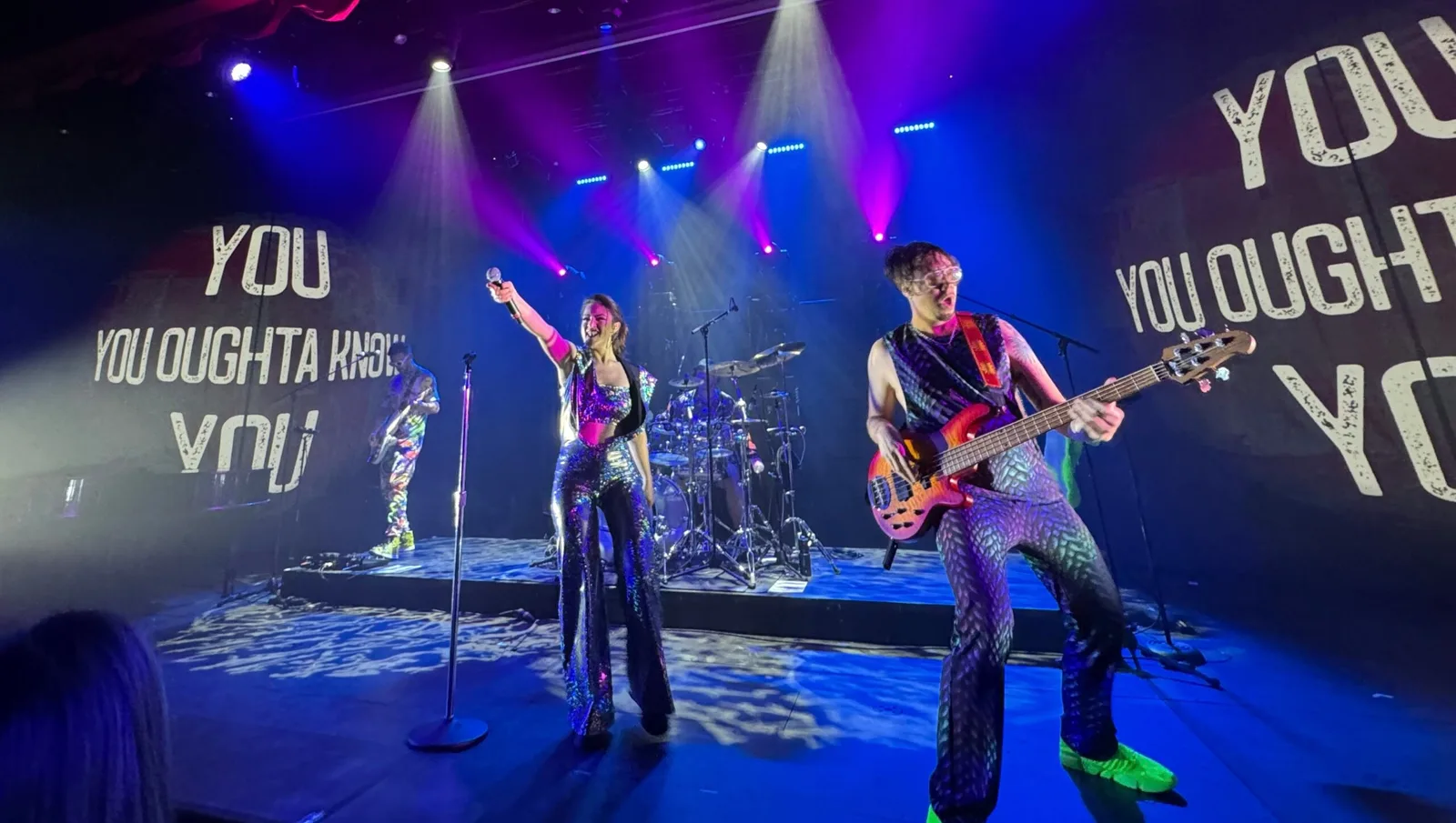 Musician playing a bass guitar on a stage with colorful lighting and partially visible text projected on the backdrop.