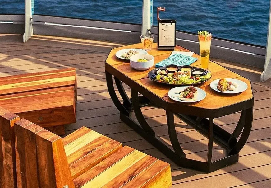 A wooden table on a boat deck set with various dishes of food, a drink, and a menu, with a view of the ocean in the background.