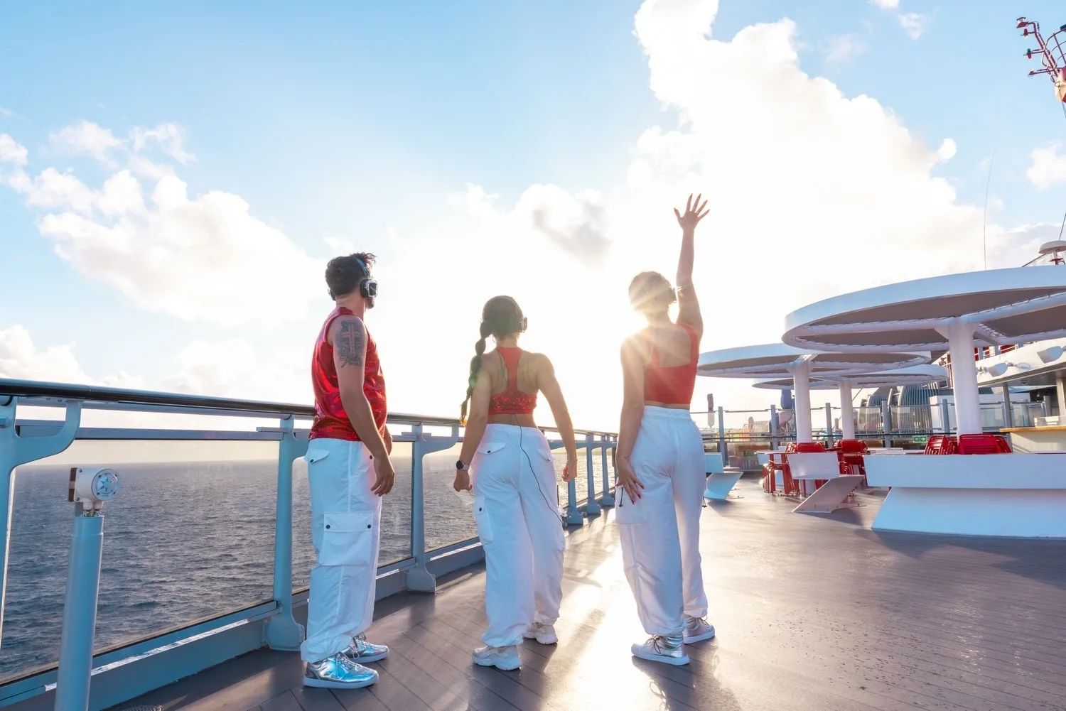 Three people wearing red tops and white pants stand on a ship deck by the railing with the ocean and sky in the background, one person raising their hand.