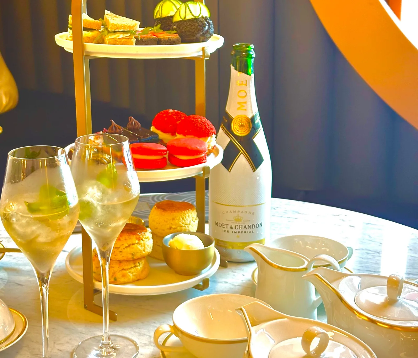 Table set with pastries, two glasses of a sparkling beverage, a bottle of Moët & Chandon champagne, and white ceramic tea service on a ship interior.