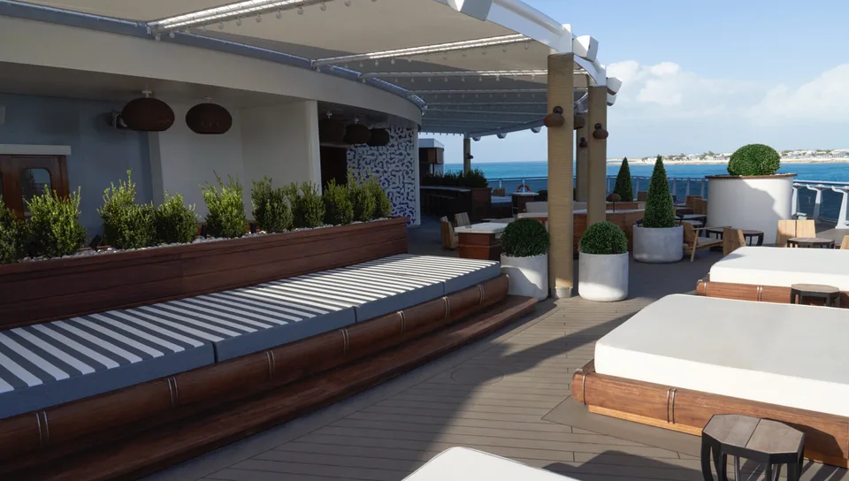 Outdoor lounge area on a ship deck with cushioned wooden daybeds, potted plants, and a view of the ocean and distant shoreline.