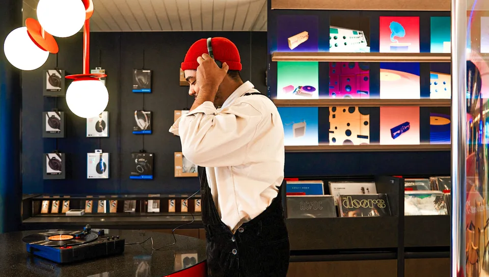 A man wearing a red cap and white shirt stands near shelves displaying brightly lit album covers and vinyl records in a music store or section.