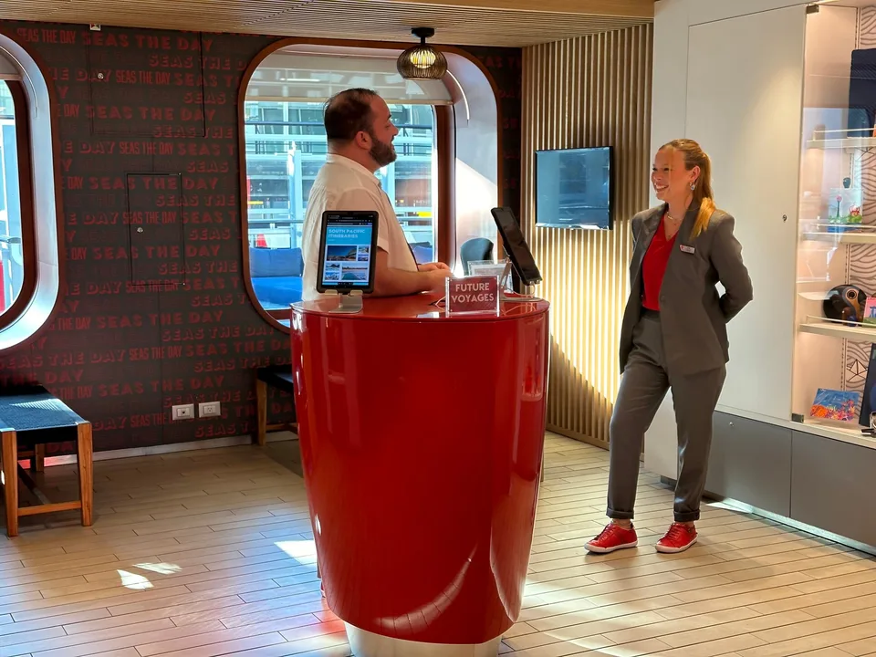 Two people stand and talk near a red reception desk inside a modern boat or ferry cabin with a window showing docked vessels outside.