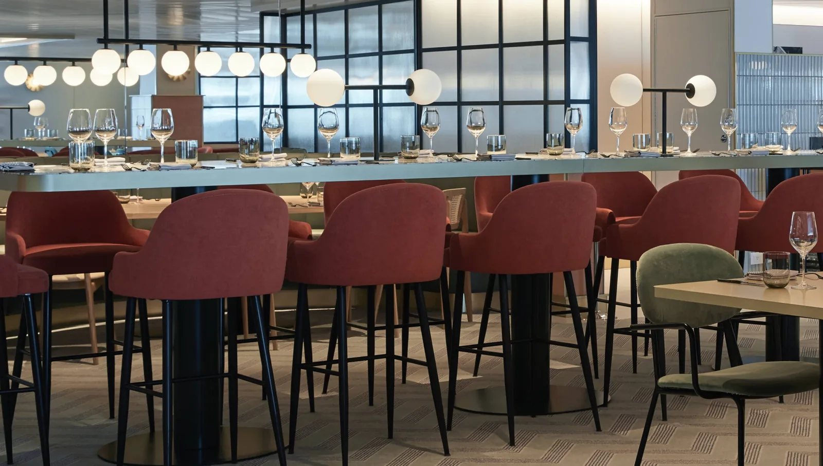Interior view of a modern bar with red upholstered high chairs, glassware on the counter, and round pendant lights above.
