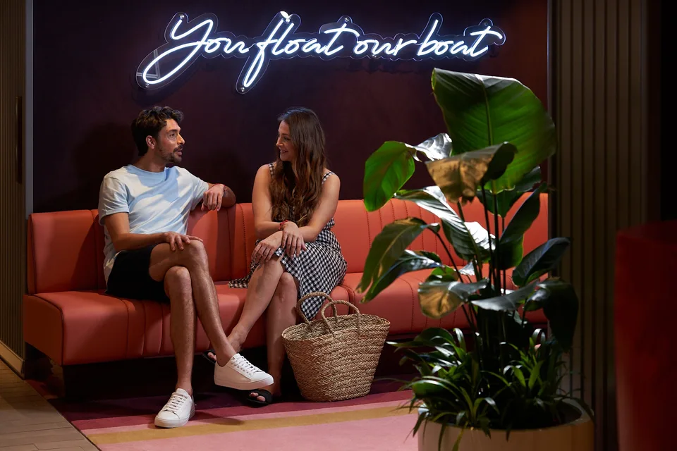 Two people sitting and talking on a red booth seat with a neon sign reading "You float our bo" in the background and a large woven bag on the floor next to them.