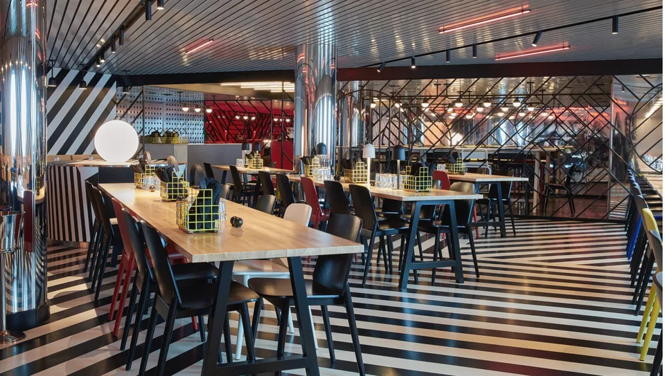 Interior of a modern onboard dining area with long wooden tables, black and red chairs, geometric decor, and polished metal columns.