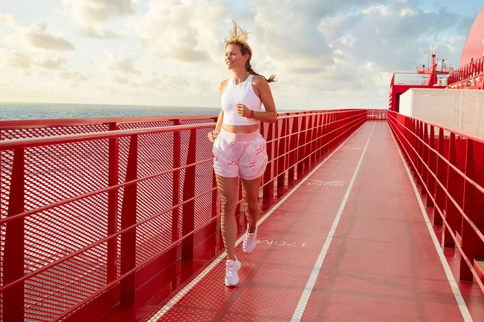 A woman wearing a white tank top and pink shorts is jogging on a red outdoor deck with railings, with the ocean and cloudy sky in the background.