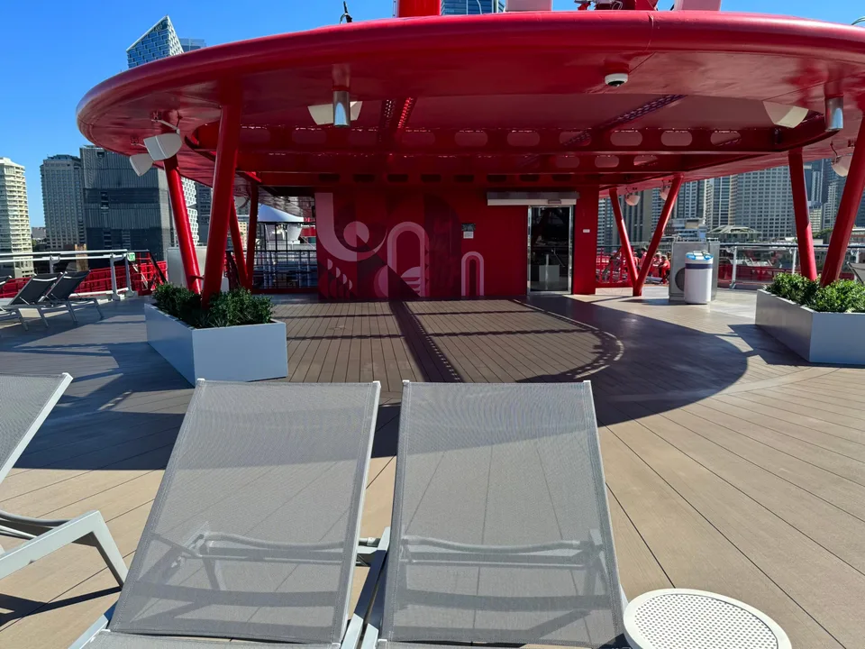 Two empty lounge chairs on the deck of a ship with a large red structure and city buildings in the background.