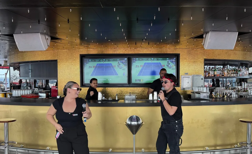 Two men sing karaoke in front of a bar with large screens displaying a tennis match, while a bartender looks on.