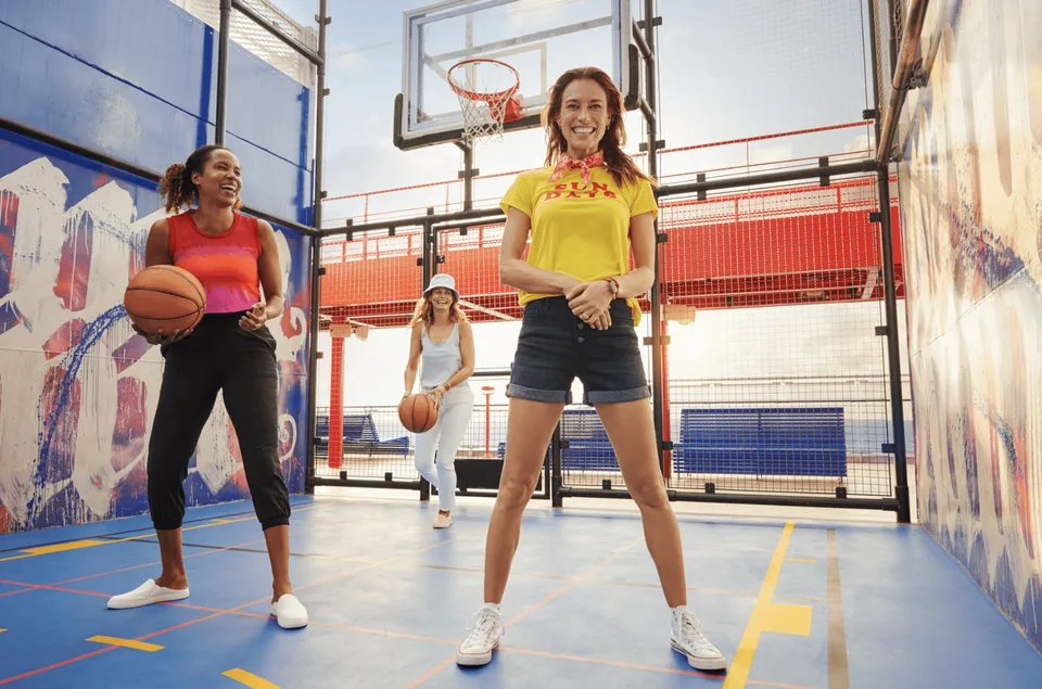 Three women standing on an indoor basketball court, two holding a basketball, with a hoop and net visible in the background.