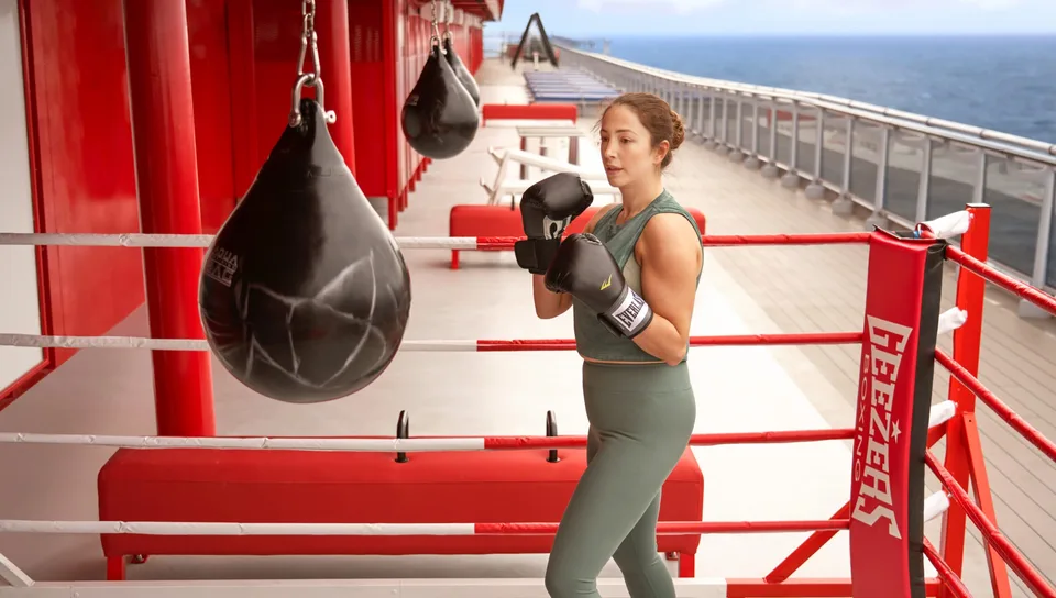 A woman wearing boxing gloves and athletic wear practices boxing in a ring on the deck of a ship with the ocean in the background.