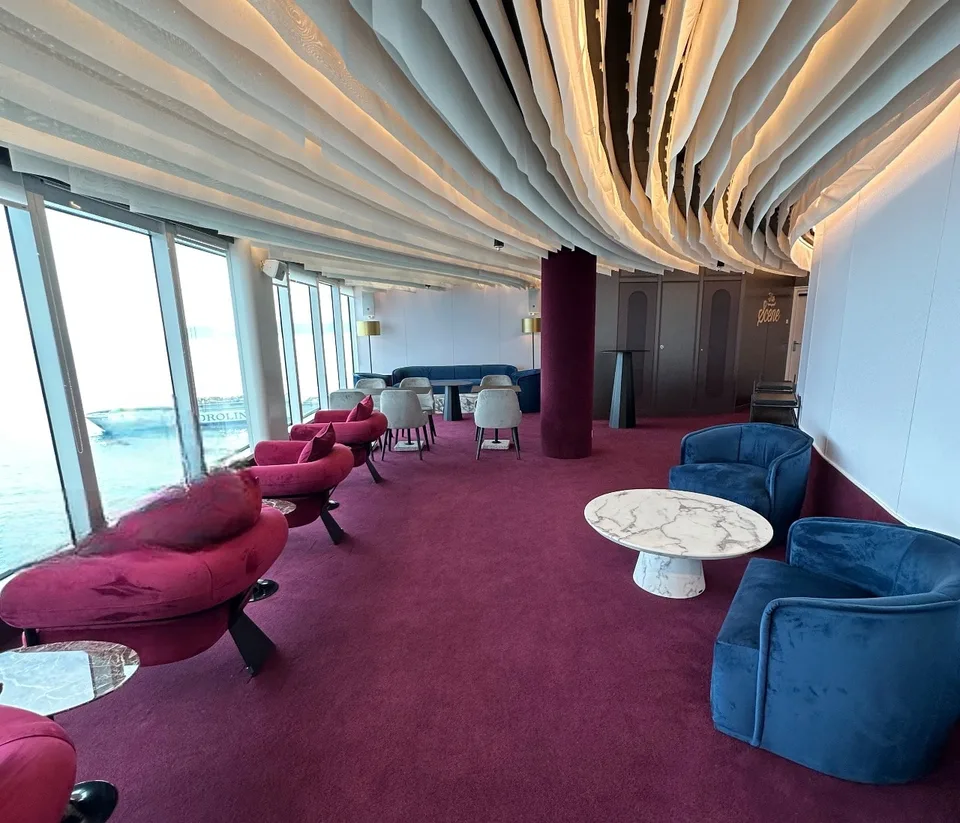 Interior lounge area on a ship with large windows, purple carpet, modern red and blue chairs, and white marble-top tables.