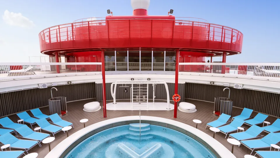 Outdoor pool area on a ship with multiple blue lounge chairs, a circular hot tub, and a red upper deck structure.