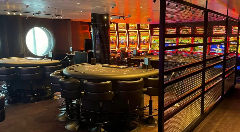 Interior of a ship's gaming or casino area featuring poker tables, cushioned stools, a large round window, and slot machines.