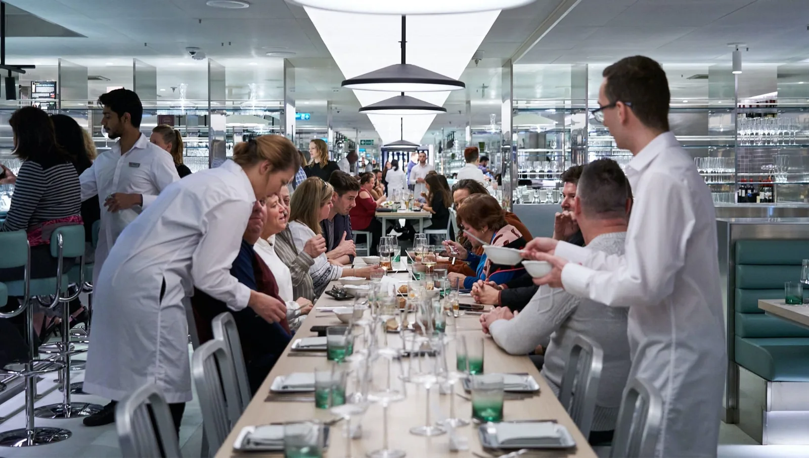 Passengers seated at long tables in a modern dining area onboard a vessel are served by staff in white uniforms.