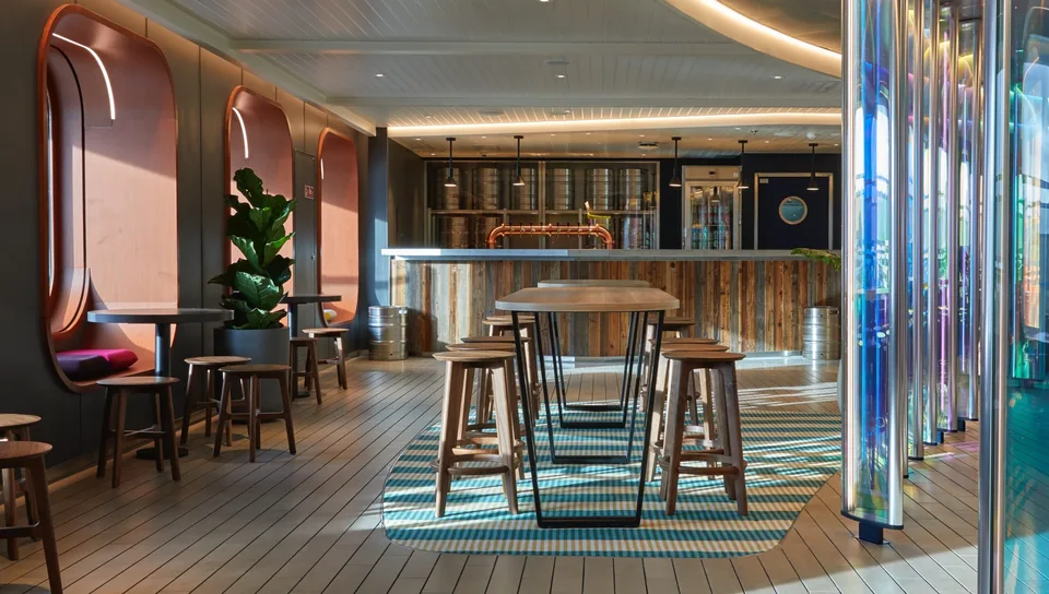 Interior image of a modern onboard dining area with wooden tables, stools, a patterned rug, and colorful vertical light panels.