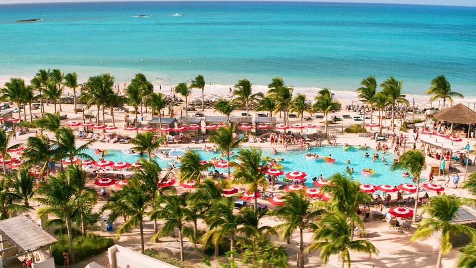 Aerial view of a tropical resort pool area with red and white umbrellas, surrounded by palm trees, people swimming and lounging, and an ocean backdrop.