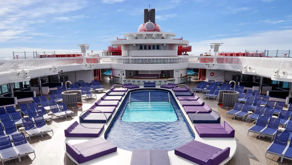 Outdoor pool area on a cruise ship with lounge chairs arranged around the pool under a partly cloudy sky.