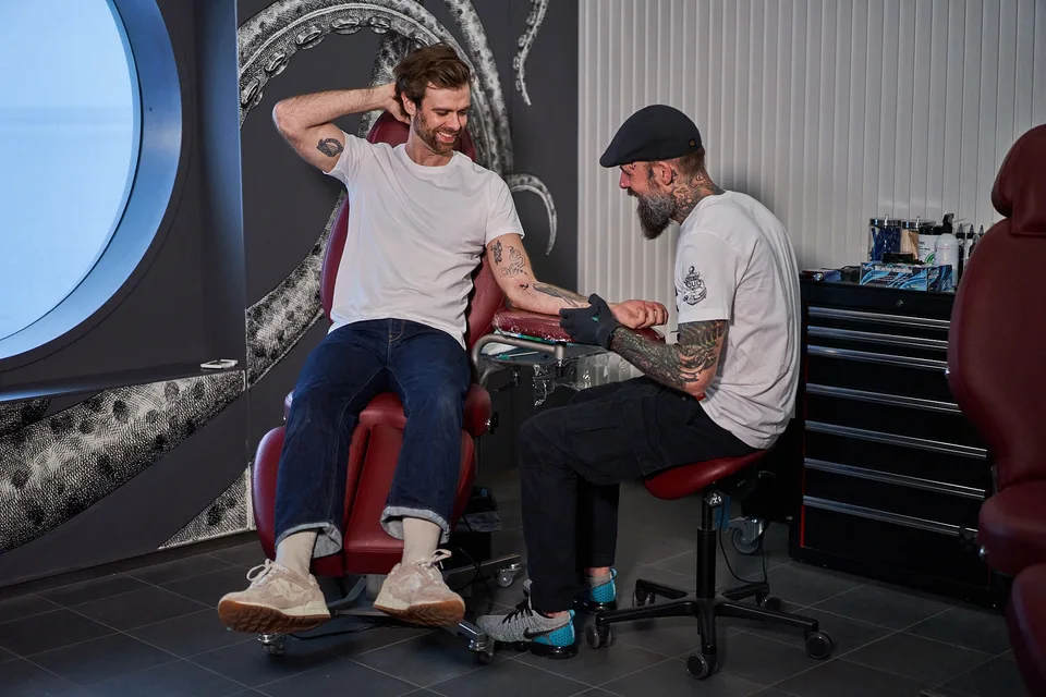 A man in a white t-shirt and rolled-up jeans is sitting on a red chair, smiling while getting a tattoo on his arm from a tattoo artist wearing a cap and gloves in a tattoo studio.