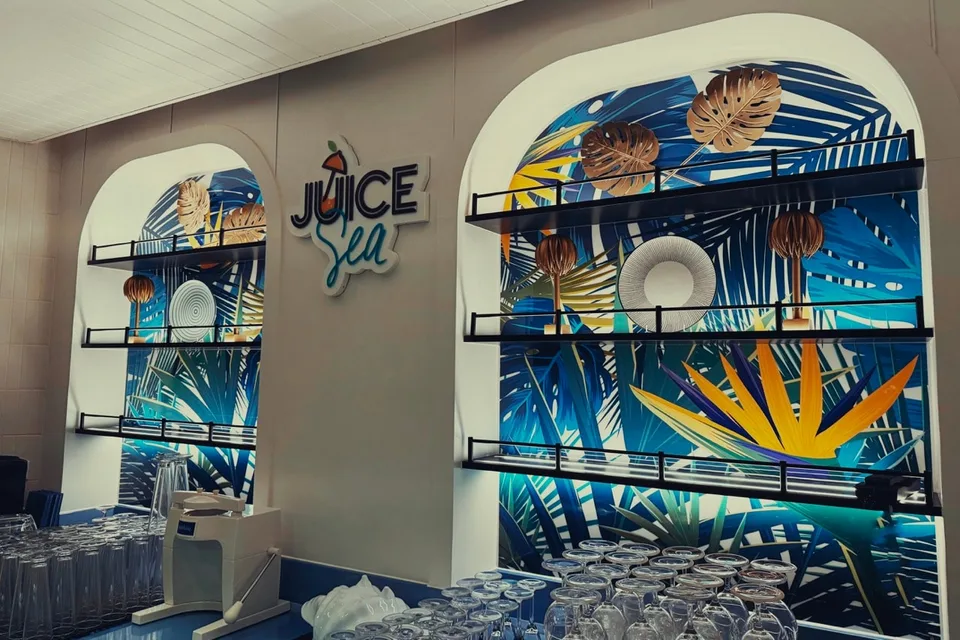 Empty glass racks mounted on a brightly colored tropical-themed wall next to part of a sign reading "Juice Sea."