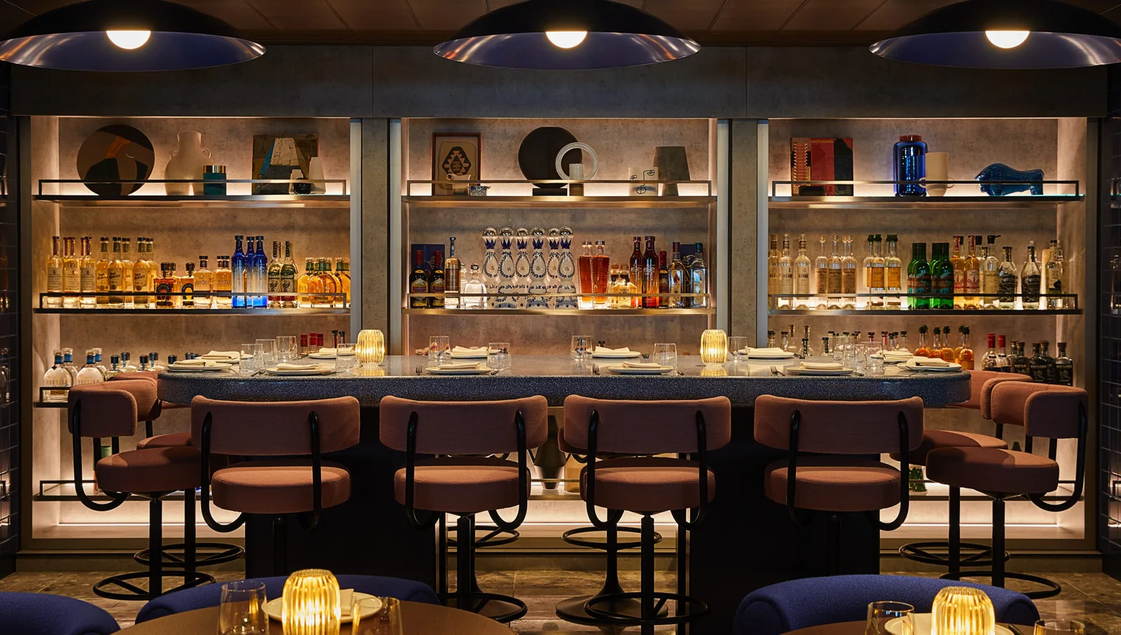 Onboard bar area with a row of padded bar stools, shelves stocked with bottles of liquor and water, and candle-lit tables.