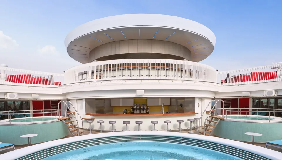 Outdoor pool area on a cruise ship with a bar, stools, and curved white architecture under a clear blue sky.