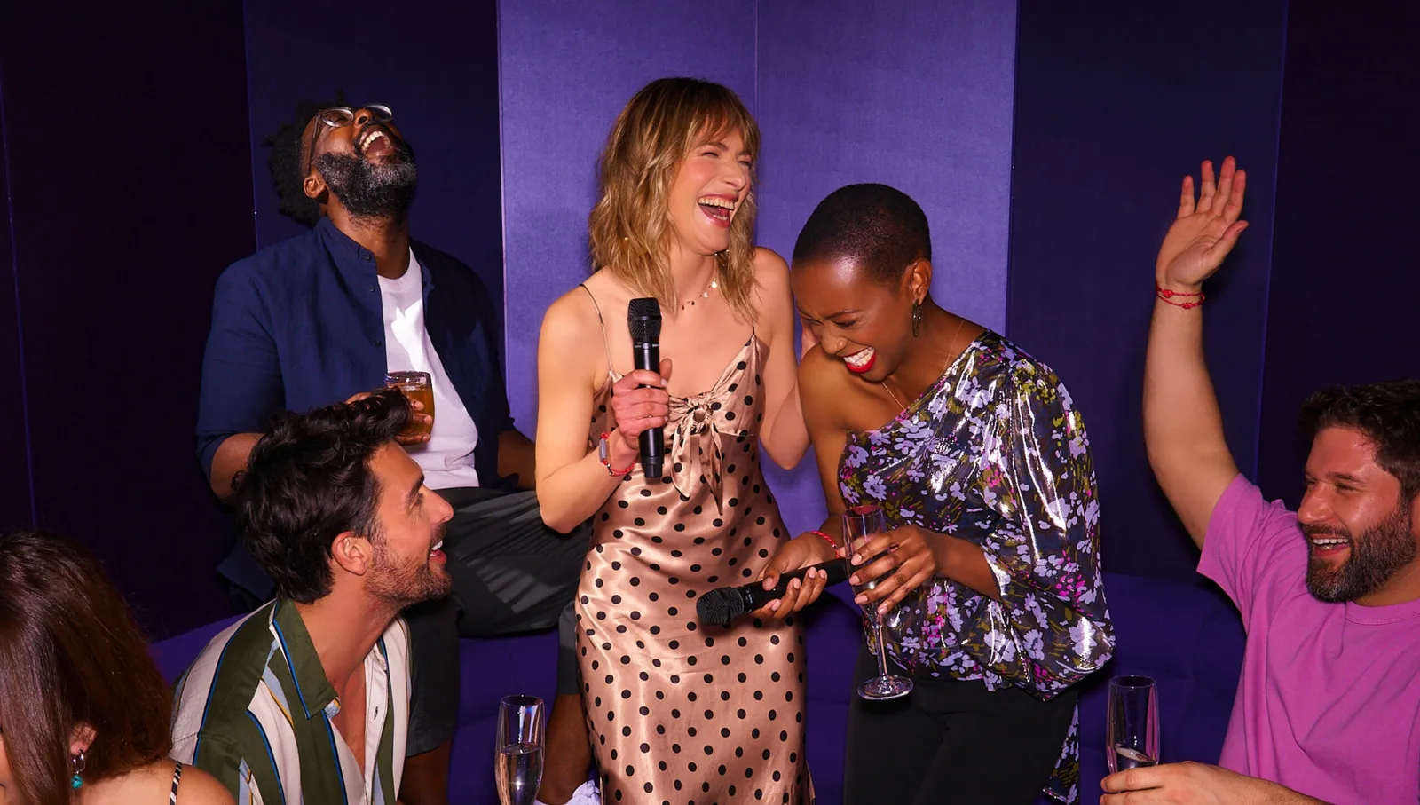 Four people laughing and holding microphones and drinks, seated and standing against a purple wall.