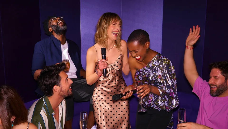 Four people laughing and holding microphones and drinks, seated and standing against a purple wall.