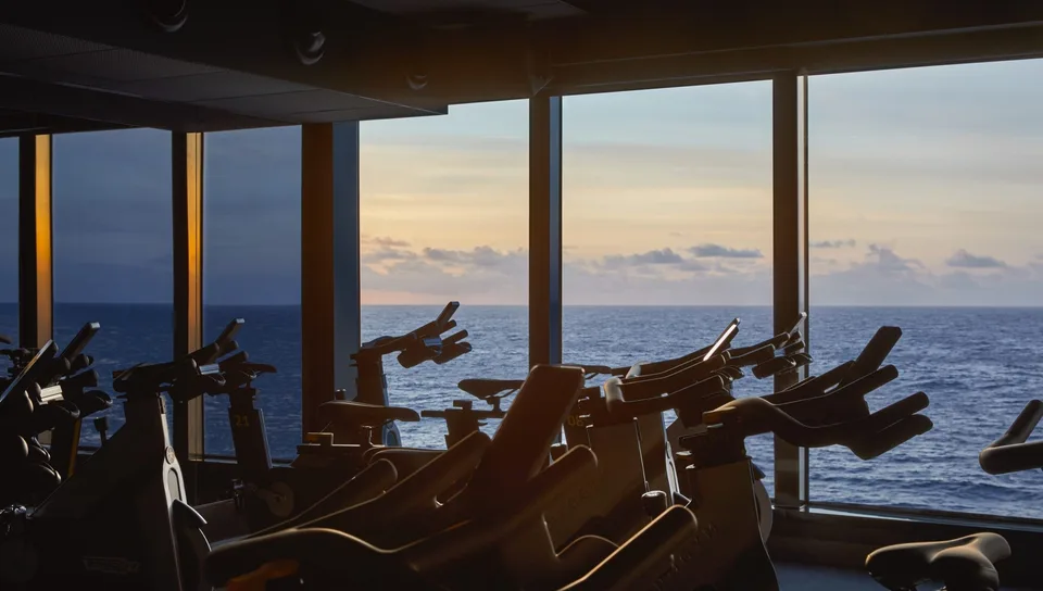 Indoor cycling bikes are arranged in a row facing large windows with a view of the ocean and a sunset sky.
