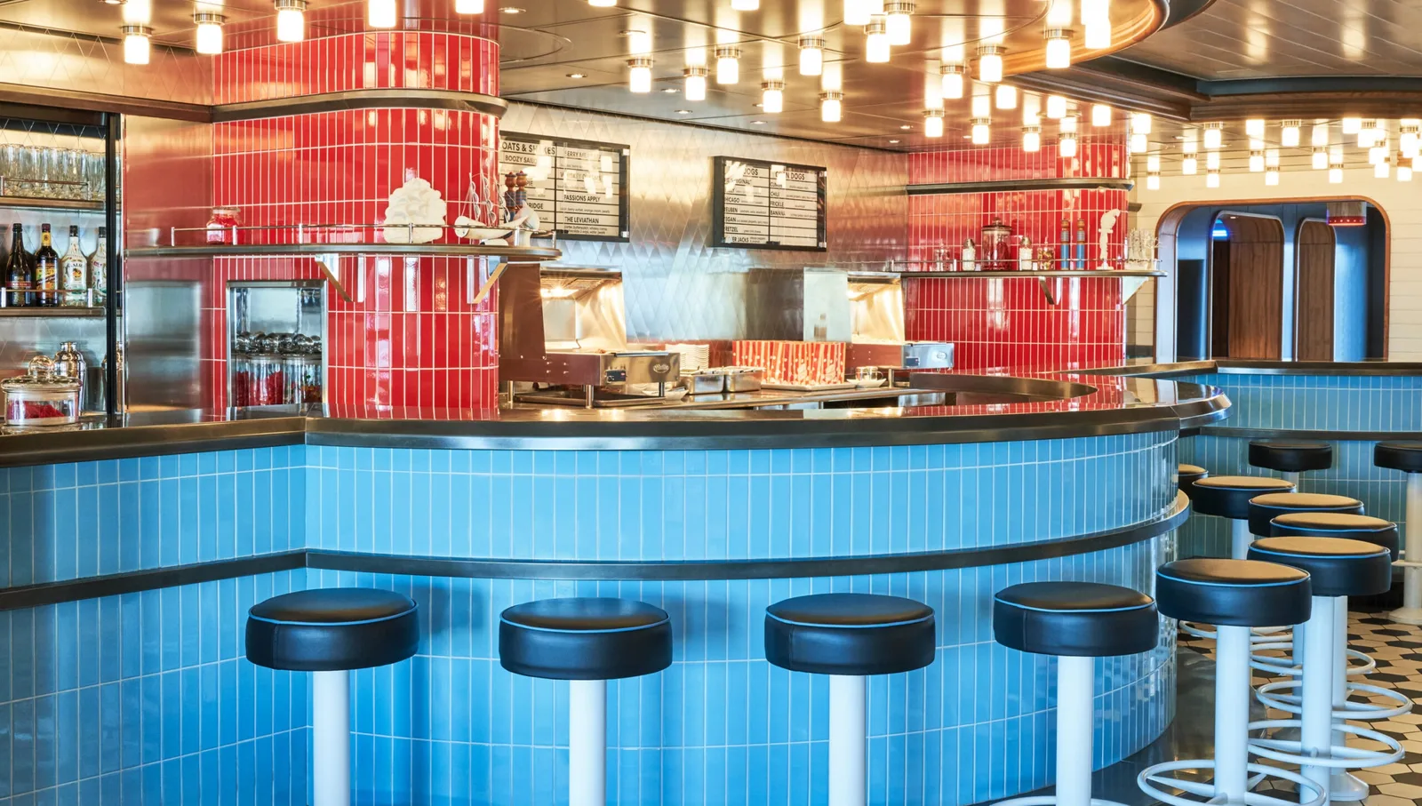 Bar area with blue-tiled curved counter, black cushioned round stools, hanging lights, and a red-tiled section with shelves and drink dispensers.