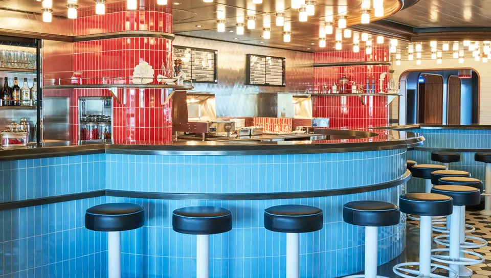Bar area with blue-tiled curved counter, black cushioned round stools, hanging lights, and a red-tiled section with shelves and drink dispensers.