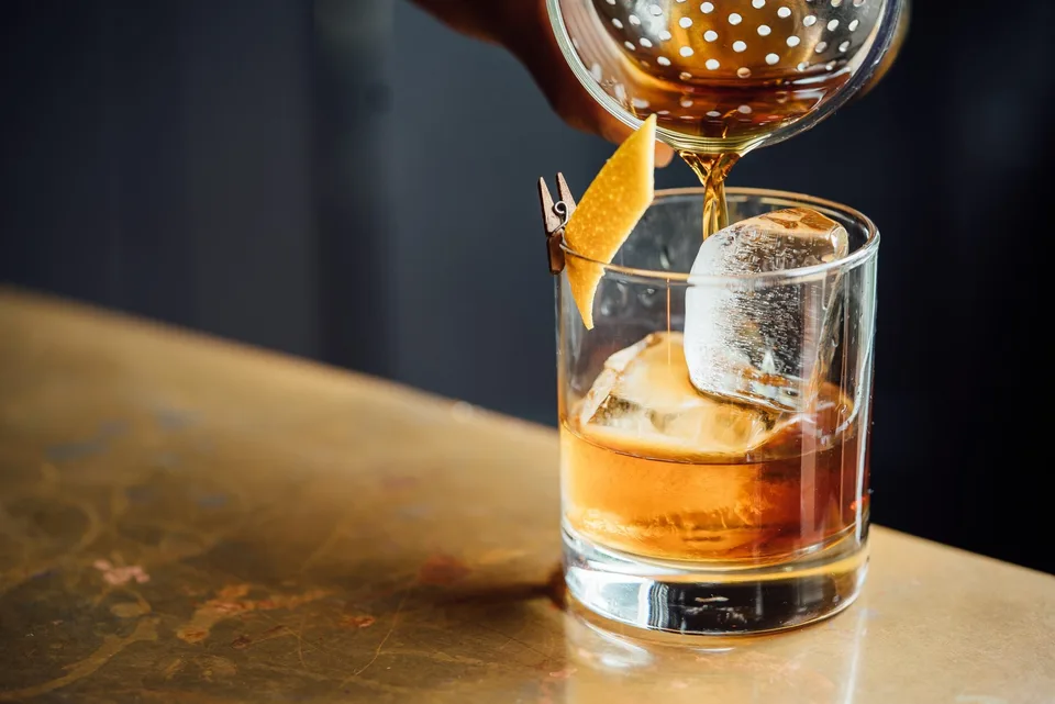 A hand is pouring a brown liquid through a strainer into a glass with a large ice cube and a lemon peel garnish.