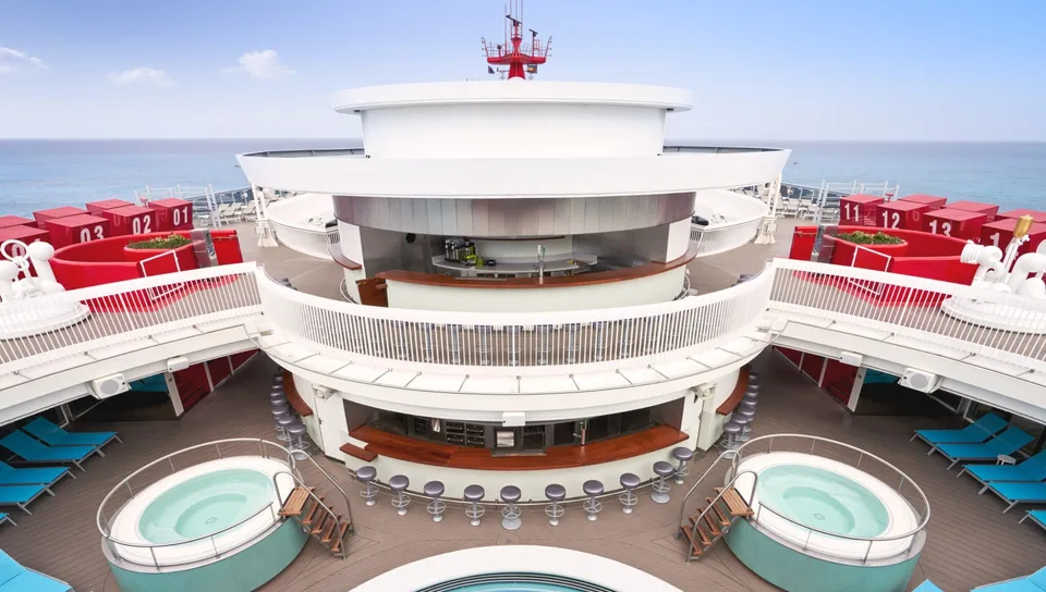 View of a circular outdoor bar area on a cruise ship deck, flanked by two hot tubs with seating around the bar and railings above.