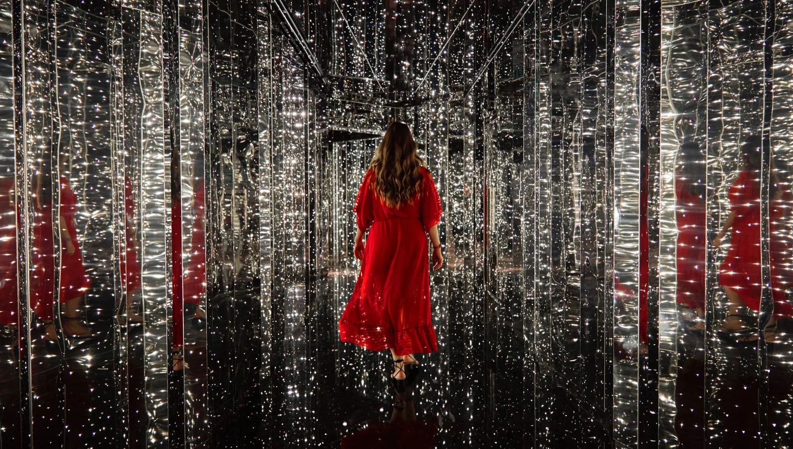 A person in a red dress walks through a hallway surrounded by mirrored walls and ceiling with numerous small lights, creating a sparkling effect.
