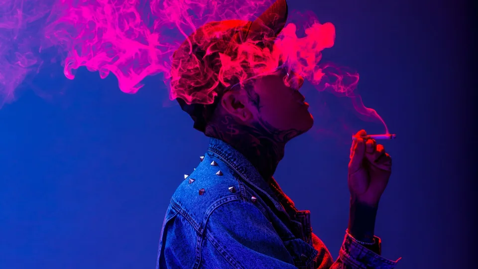A person wearing a studded denim jacket with pink smoke obscuring their face against a blue background.