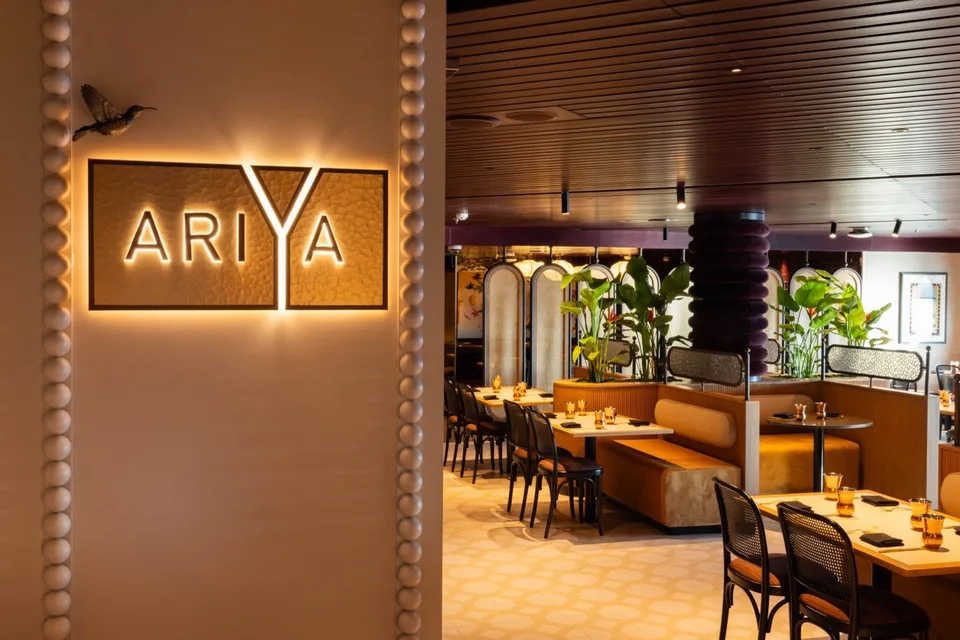 Interior view of a modern onboard dining area with wooden tables, cushioned booths, black chairs, and decorative plants.