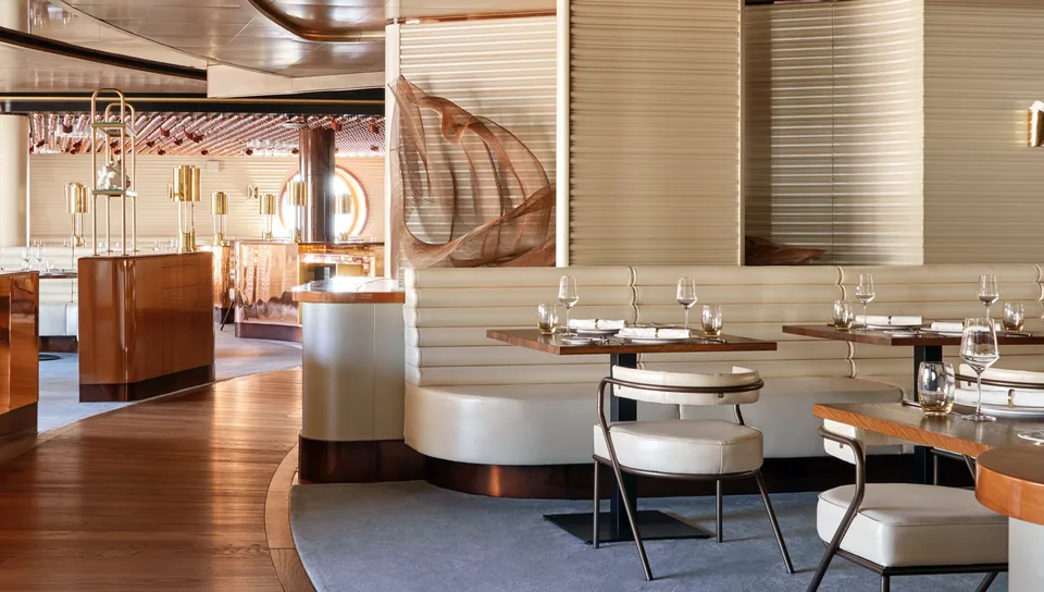 Interior view of a modern onboard dining area with wooden floors, white cushioned seating, and decorative elements including a saxophone and hanging glassware.