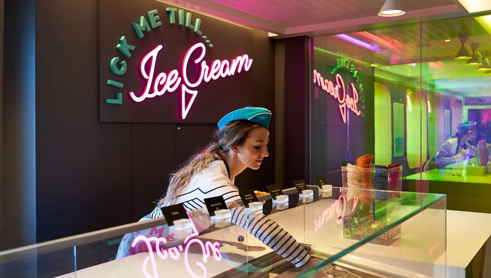 A woman in a striped shirt and blue hat is serving ice cream behind a glass counter with colorful neon lights reflected in the background.