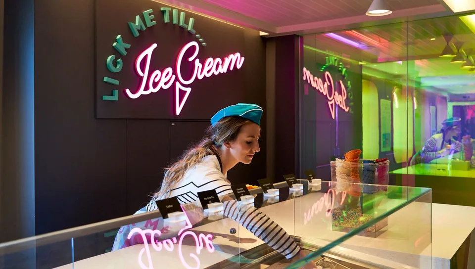 A woman in a striped shirt and blue hat is serving ice cream behind a glass counter with colorful neon lights reflected in the background.